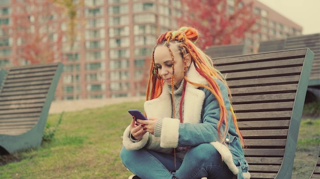 Young woman with dreadlocks in warm clothes browsing mobile phone sitting on wooden sunbed in city park Carefree female with colourful hairstyle using smartphone on walk in cloudy weather