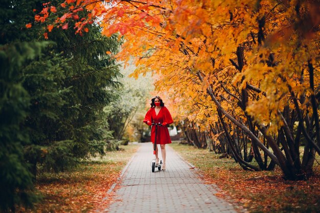 Young woman with electric scooter in red dress at the autumn city park