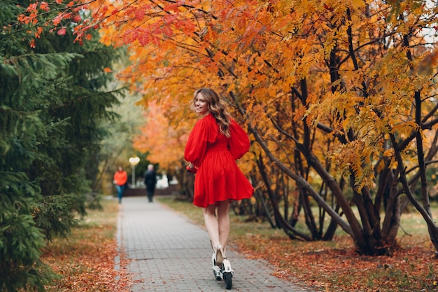 Young woman with electric scooter in red dress at the autumn city park