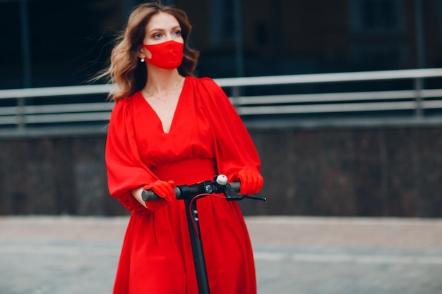 Young woman with electric scooter in red dress and gloves with face mask at the city New normal fashion and coronavirus COVID protection concept