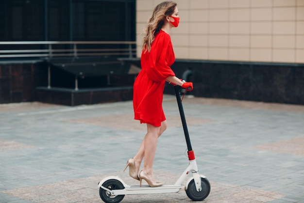 Young woman with electric scooter in red dress and gloves with face mask at the city New normal fashion and coronavirus COVID protection concept