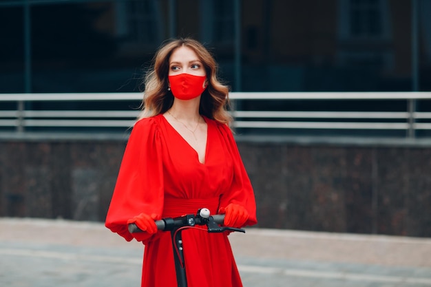 Young woman with electric scooter in red dress and gloves with medical face mask at the city