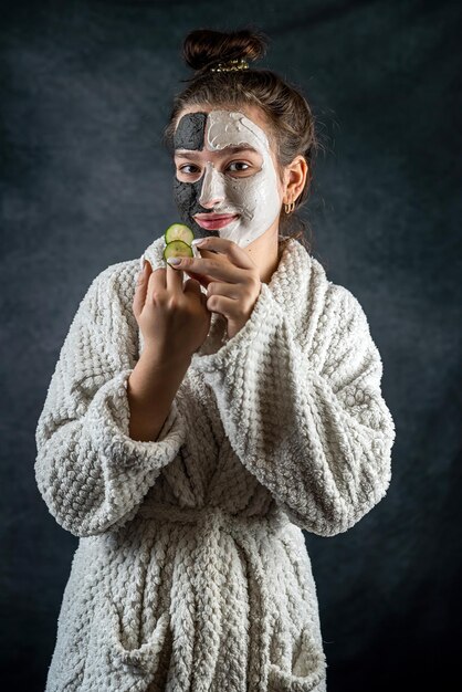 Young woman with facial clay black and white mask holding cucumber slice isolated on dark