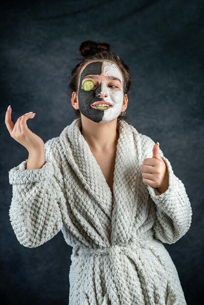 Young woman with facial clay black and white mask holding cucumber slice isolated on dark