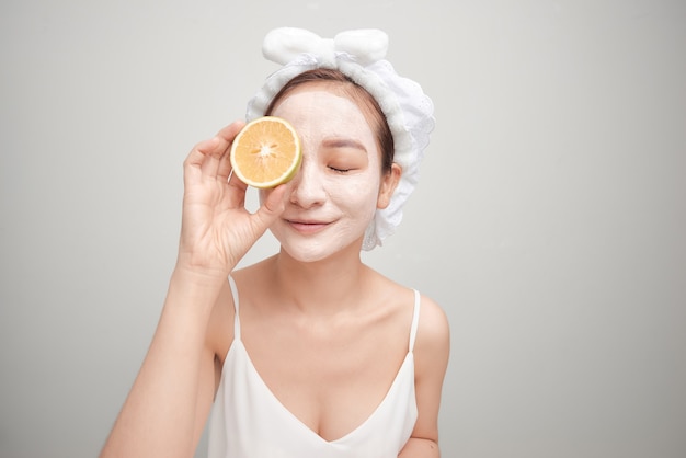 Young woman with facial clay mask holding orange fruit slice covering eyes