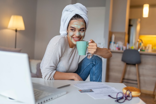 Young woman with facial mask applied drinking coffee while using laptop.