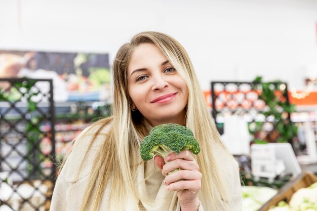 Young woman with fresh broccoli at the grocery store. Smiling blonde in a beige coat. Healthy food, vitamins and vegetarianism. Close-up.