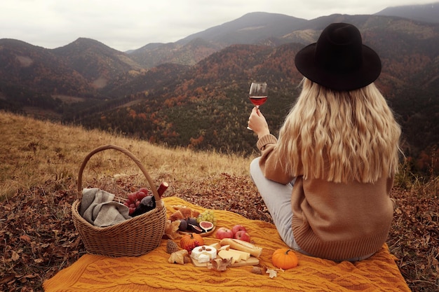 Young woman with glass of wine having picnic in mountains on autumn day back view