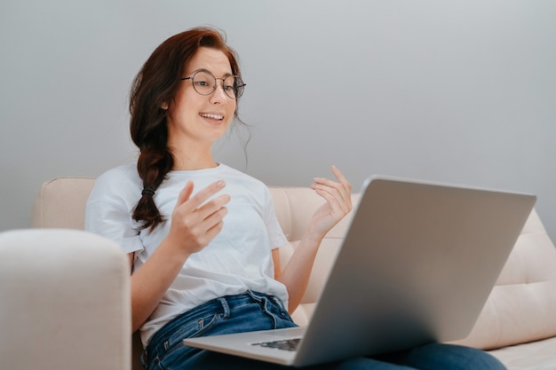 Young woman with glasses communicates on the internet sitting on a bright sofa with a laptop at home...