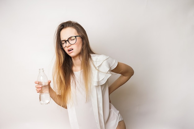 A young woman with glasses with long hair feels the pain in her back, holds the bottle in her hand, closes her eyes in pain