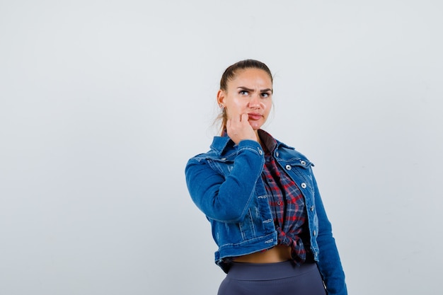 Young woman with hand near mouth in checkered shirt, jean jacket and looking puzzled .