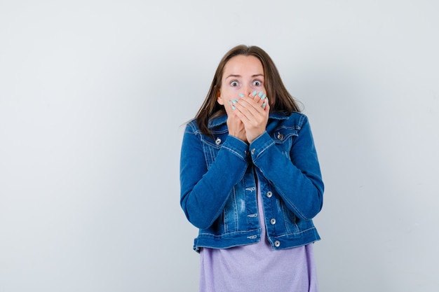 Young woman with hands on mouth in denim jacket and looking frightened. front view.