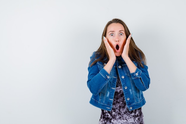 Young woman with hands near face in denim jacket and looking shocked , front view.