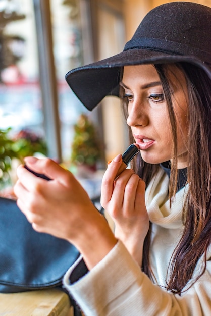 Young woman with a hat puts lipstick on lips