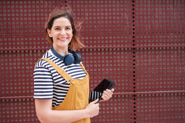 Young woman with headphones and tablet dressed in yellow dungarees on a red background outdoors Concept Lifestyle youth trends