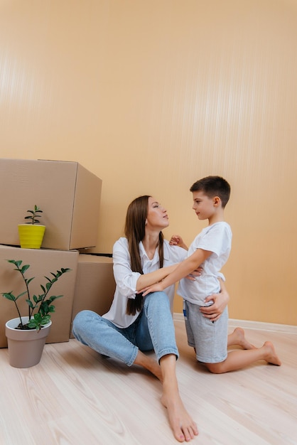 A young woman with her son is sitting in front of the boxes and rejoicing at the housewarming after moving in. Housewarming, delivery and freight transportation, purchase of real estate.