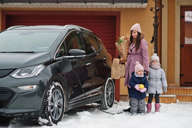 Young woman with kids hold eco bags and charging electric car in the yard of her house