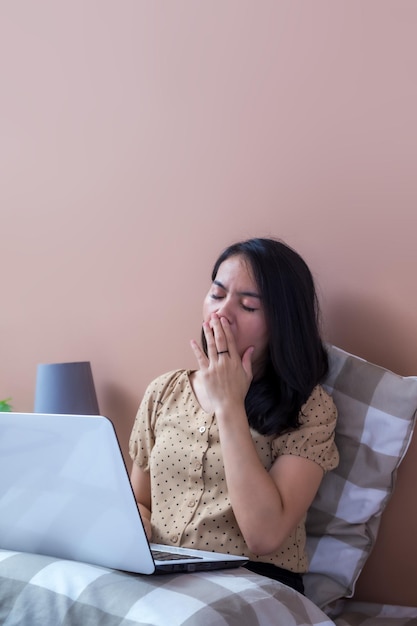 Young woman with laptop in bed yawning with hands covering mouth