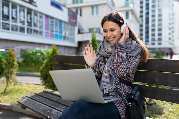 Young woman with a laptop on her knees in headphones outside smiling with delight and throwing up