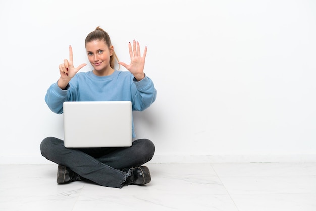 Young woman with a laptop sitting on the floor counting seven with fingers