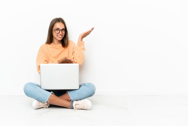 Young woman with a laptop sitting the floor extending hands to the side for inviting to come