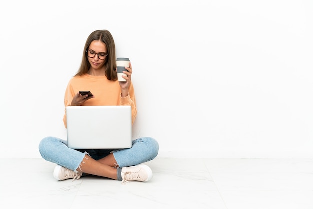 Young woman with a laptop sitting on the floor holding coffee to take away and a mobile