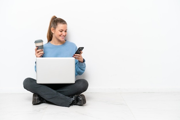 Young woman with a laptop sitting on the floor holding coffee to take away and a mobile