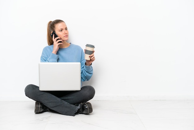 Young woman with a laptop sitting on the floor holding coffee to take away and a mobile