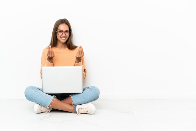 Young woman with a laptop sitting on the floor making money gesture