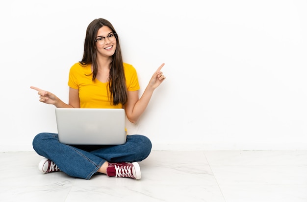 Young woman with a laptop sitting on the floor pointing finger to the laterals and happy