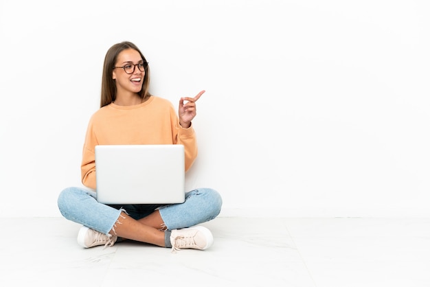 Young woman with a laptop sitting on the floor pointing finger to the side and presenting a product