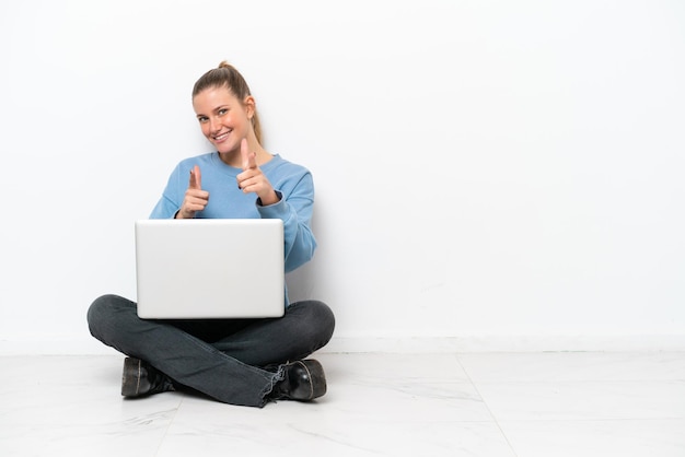 Young woman with a laptop sitting on the floor pointing to the front and smiling