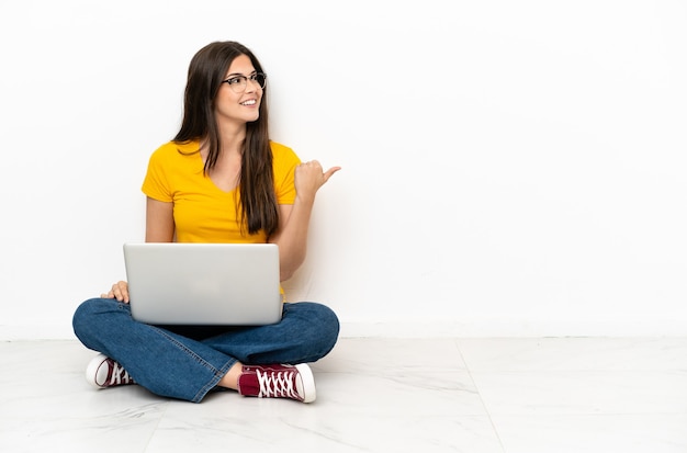 Young woman with a laptop sitting on the floor pointing to the side to present a product