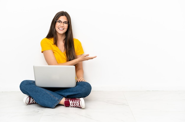 Young woman with a laptop sitting on the floor presenting an idea while looking smiling towards