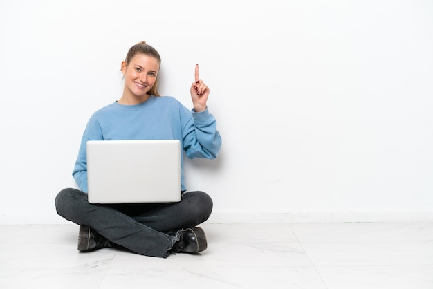 Young woman with a laptop sitting on the floor showing and lifting a finger in sign of the best