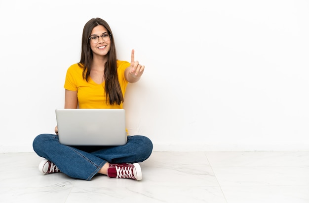 Young woman with a laptop sitting on the floor showing and lifting a finger