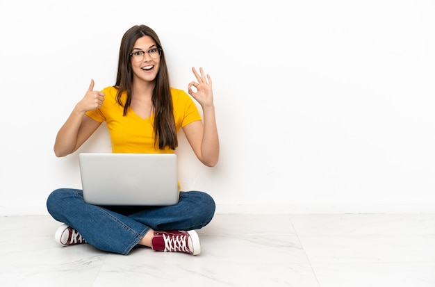 Young woman with a laptop sitting on the floor showing ok sign and thumb up gesture