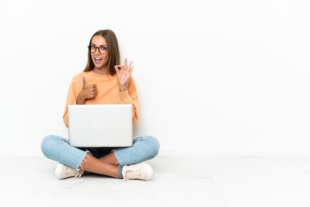 Young woman with a laptop sitting on the floor showing ok sign and thumb up gesture