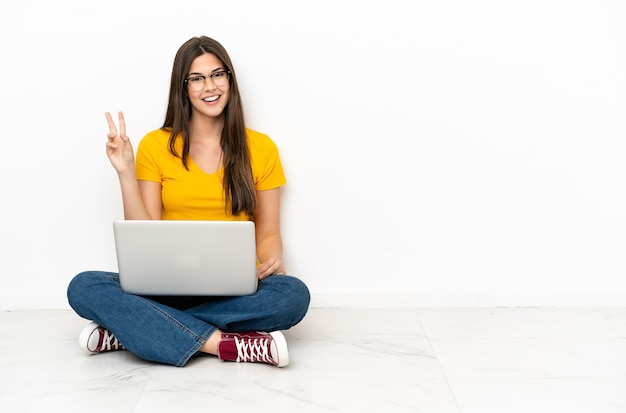 Young woman with a laptop sitting on the floor smiling and showing victory sign