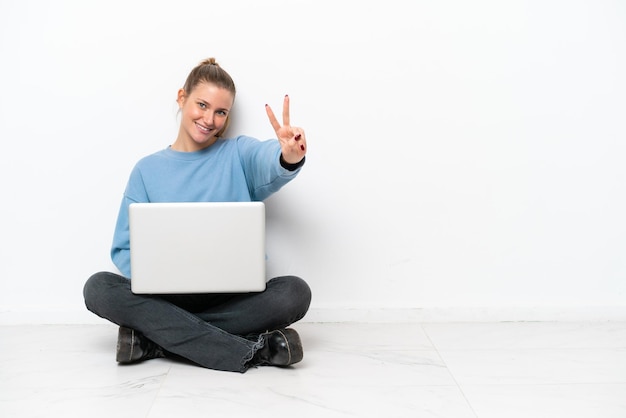 Young woman with a laptop sitting on the floor smiling and showing victory sign