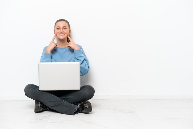 Young woman with a laptop sitting on the floor smiling with a happy and pleasant expression