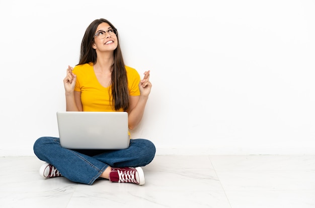 Young woman with a laptop sitting on the floor with fingers crossing