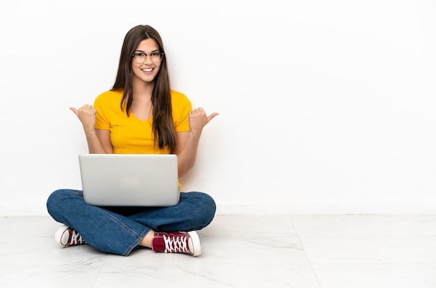 Young woman with a laptop sitting on the floor with thumbs up gesture and smiling