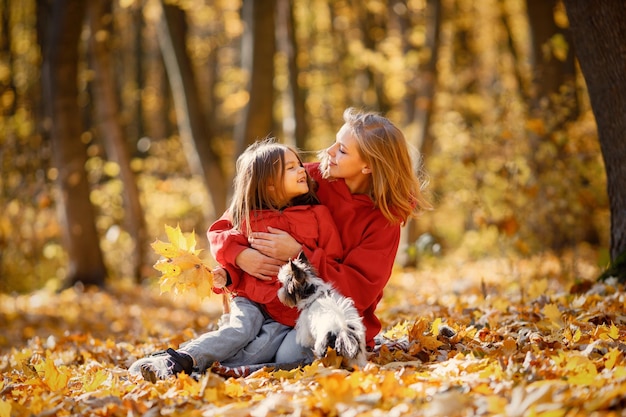 Young woman with little girl sitting on a blanket in autumn forest. Blonde woman play with her daughter and holding yorkshire terrier dog. Mother and daughter wearing jeans and red jackets.