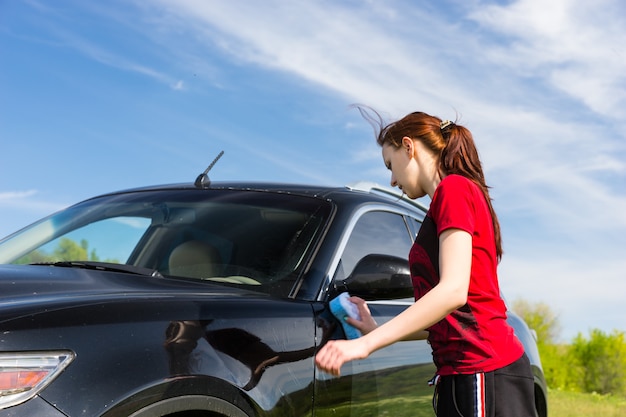 Young Woman with Long Auburn Hair and Wearing Red Shirt Washing Black Luxury Vehicle with Sponge in Green Field on Sunny Day