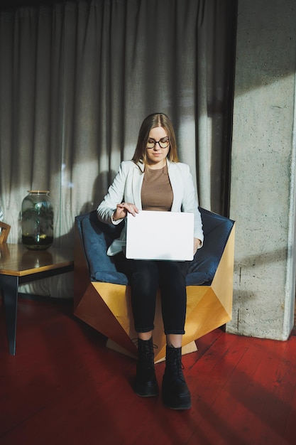 A young woman with long blond hair in stylish clothes is sitting in a chair and working on a laptop Work in a modern office with large windows