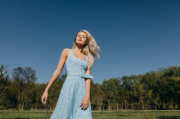 Young woman with long blonde hair wearing a blue dress posing on blue sky background outdoors