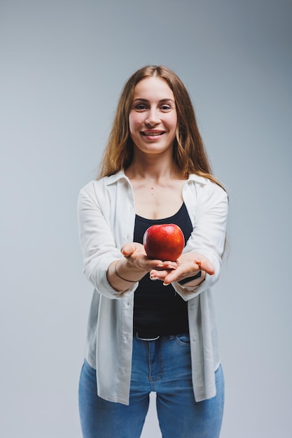 A young woman with long brunette hair dressed in a white shirt and jeans holds a fresh red apple in her hands Healthy food concept White background