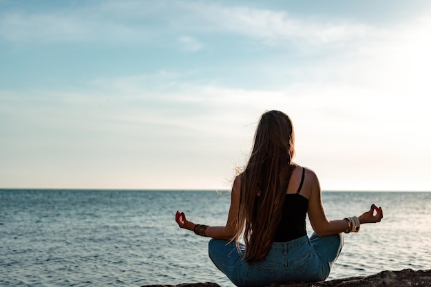 Young woman with long hair in black swimsuit and boho style braclets practicing outdoors on yoga mat by the sea on a sunset Women's yoga fitness routine Healthy lifestyle harmony and meditation