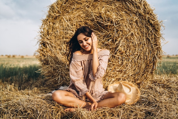 A young woman with long hair and in a dress sits near a hay bale. Woman posing smiling and looking at camera